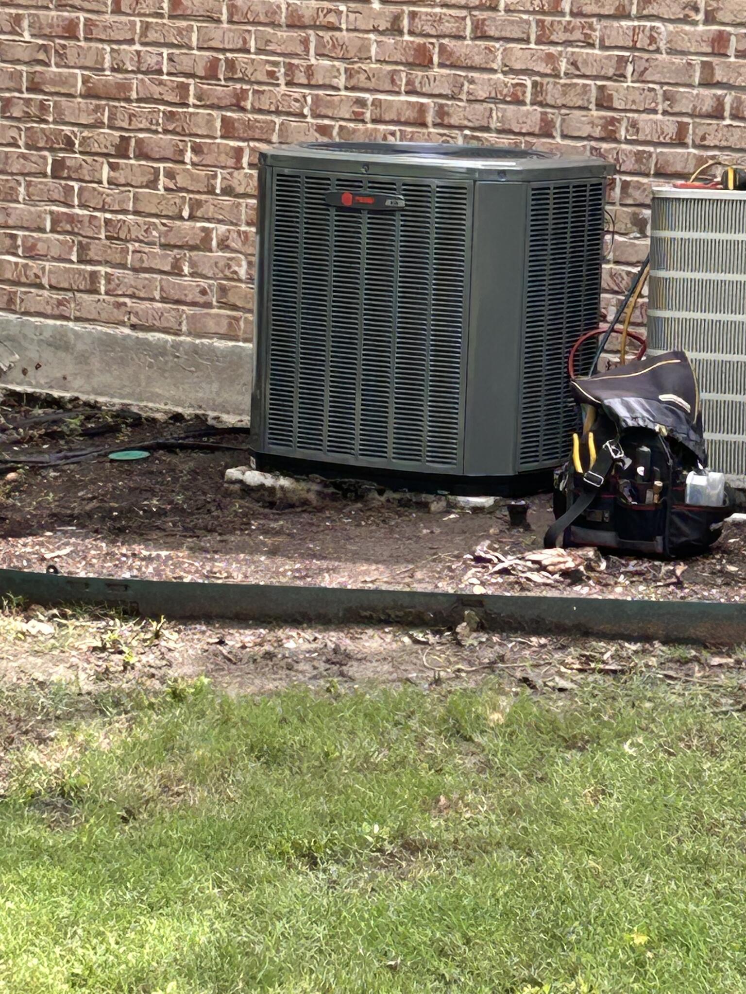 Newly installed Trane air conditioning condenser unit against a brick house exterior, with a technician's tool bag beside it